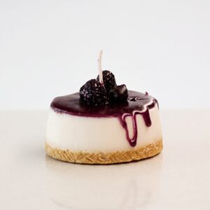 Vela decorativa artesanal hiperrealista en forma de mini cheesecake con sirope de frutos del bosque morado goteando y moras