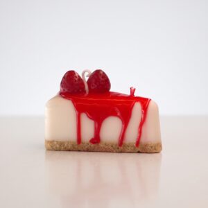 Vela decorativa artesanal hiperrealista en forma de porción de cheesecake con sirope rojo goteando y dos fresas