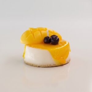 Vela decorativa artesanal hiperrealista en forma de postre o cheesecake de mango con sirope amarillo brillante goteando, trozo de mango tallado y arándanos de cera